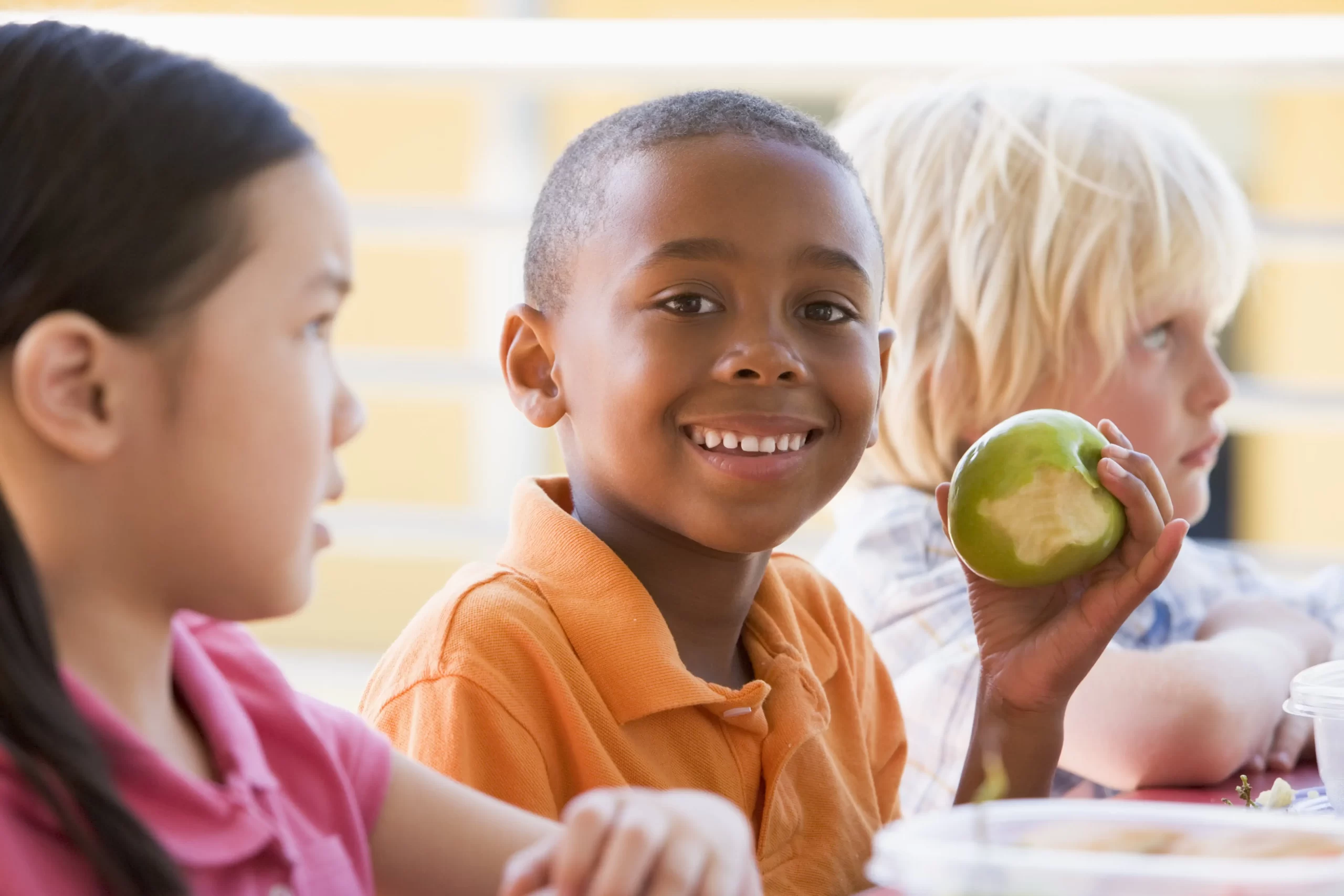 shutterstock_77073568-min Criança a sorrir e a segurar uma maçã verde mordida sentada à mesa com outras crianças